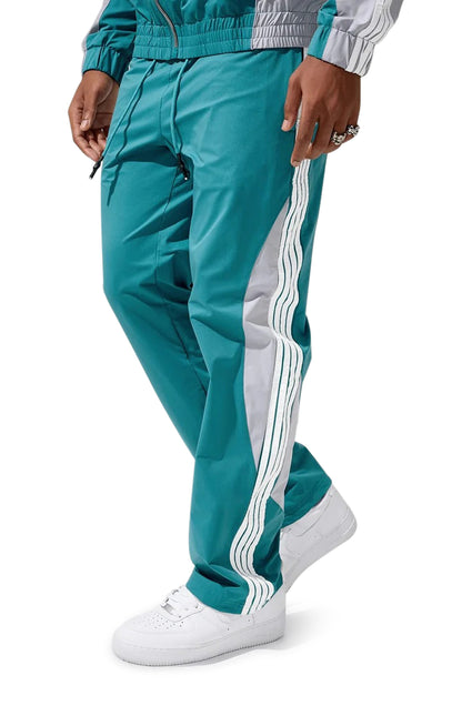 Jordan Craig teal track pants featuring silver side panels and white triple stripes, worn with white sneakers.