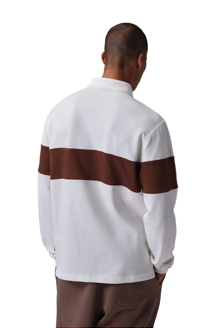 Back view of a man wearing a Nike Jordan Brooklyn rugby shirt in white and bronze with brown pants on white background.