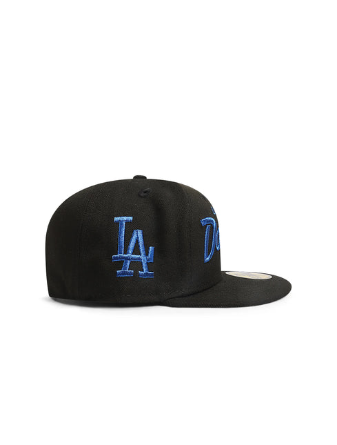 Side view of a black fitted baseball cap with a blue LA logo embroidered on the side.